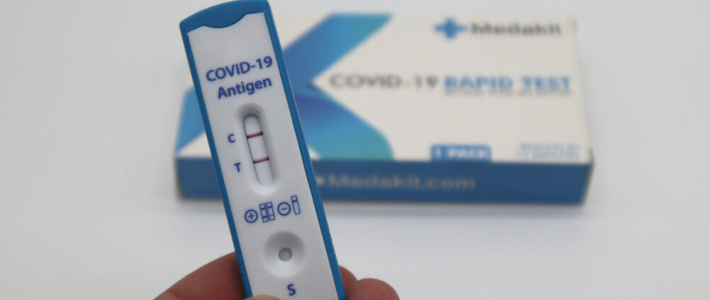 Eine Hand hält einen positiven Covid-Schnelltest. Im Hintergrund liegt die Packung des Tests.