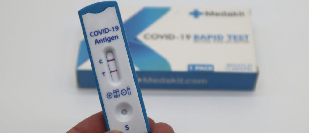 Eine Hand hält einen positiven Covid-Schnelltest. Im Hintergrund liegt die Packung des Tests.