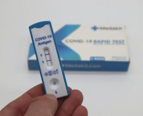 Eine Hand hält einen positiven Covid-Schnelltest. Im Hintergrund liegt die Packung des Tests.