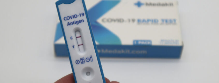 Eine Hand hält einen positiven Covid-Schnelltest. Im Hintergrund liegt die Packung des Tests.